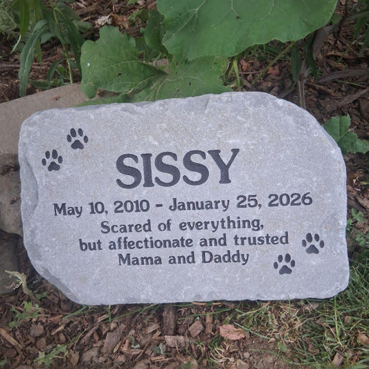 A personalized pet memorial stone engraved with the name 'SISSY', dates, and paw prints, placed outdoors on the ground among grass, mulch, and green leaves.