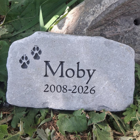 A personalized pet memorial garden stone engraved with the name 'Moby', the dates '2008-2026', and two black paw prints, placed outdoors among green leaves and grass.