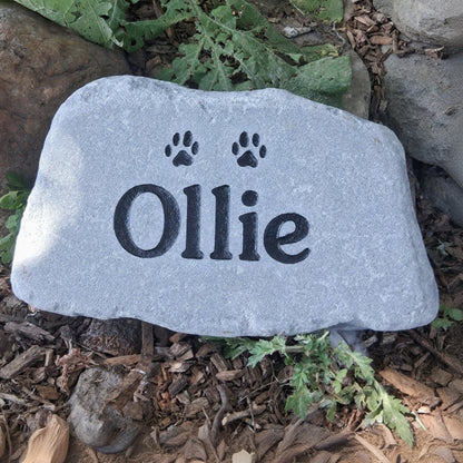 A custom pet memorial garden stone engraved with the name 'Ollie' and two paw prints, placed on a bed of mulch and green plants outdoors.