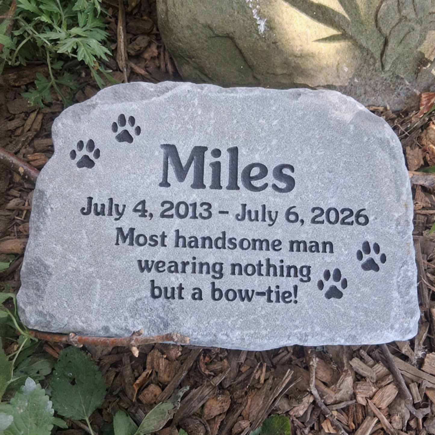 A personalized pet memorial stone engraved with the name 'Miles', dates, a quote, and paw prints, placed outdoors on mulch and greenery.