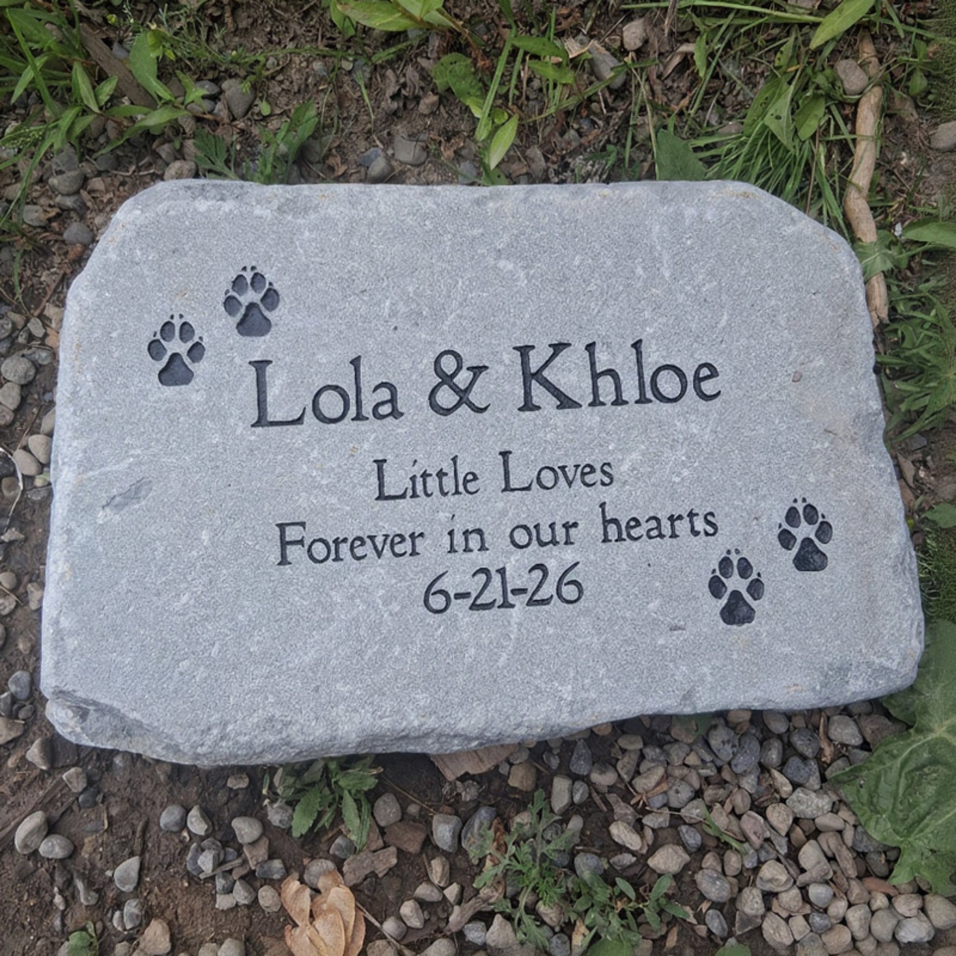 A personalized gray stone pet memorial plaque engraved with the names 'Lola & Khloe', 'Little Loves', 'Forever in our hearts', and the date '6-21-26', along with black paw print designs. The stone is photographed lying on a natural ground surface of dirt, grass, and small gray pebbles.