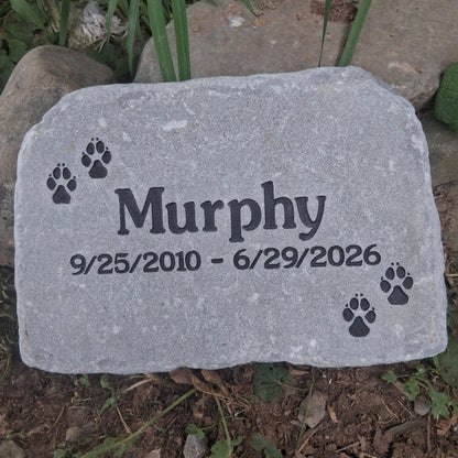 A personalized gray pet memorial garden stone with the name 'Murphy', dates '9/25/2010 - 6/29/2026', and black paw prints, shown outdoors on soil and grass.