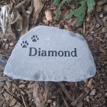 A gray personalized pet memorial garden stone engraved with the name 'Diamond' and paw prints, placed on a bed of brown wood mulch.