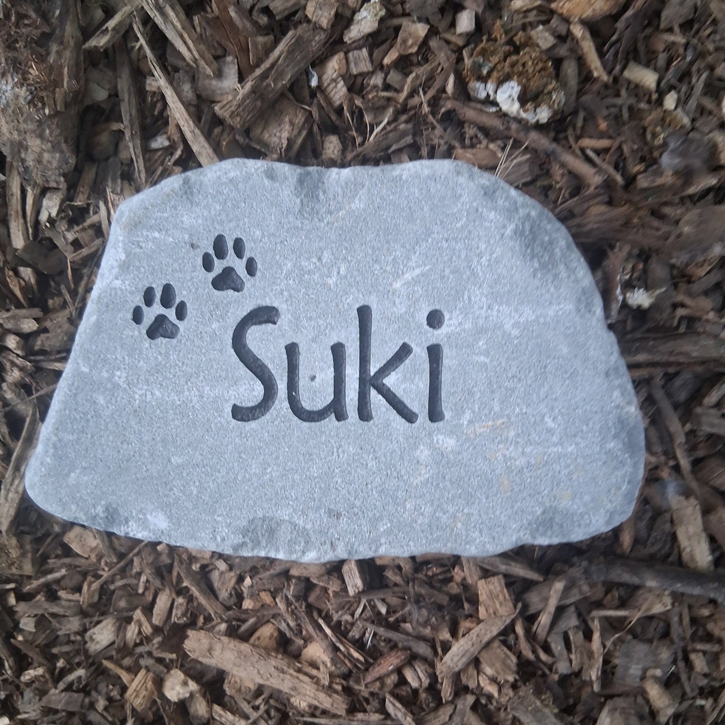 A custom pet memorial garden stone engraved with the name 'Suki' and paw prints, resting on a bed of brown wood mulch.