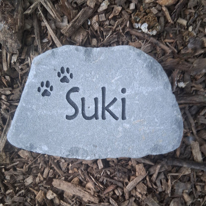A custom pet memorial garden stone engraved with the name 'Suki' and paw prints, resting on a bed of brown wood mulch.