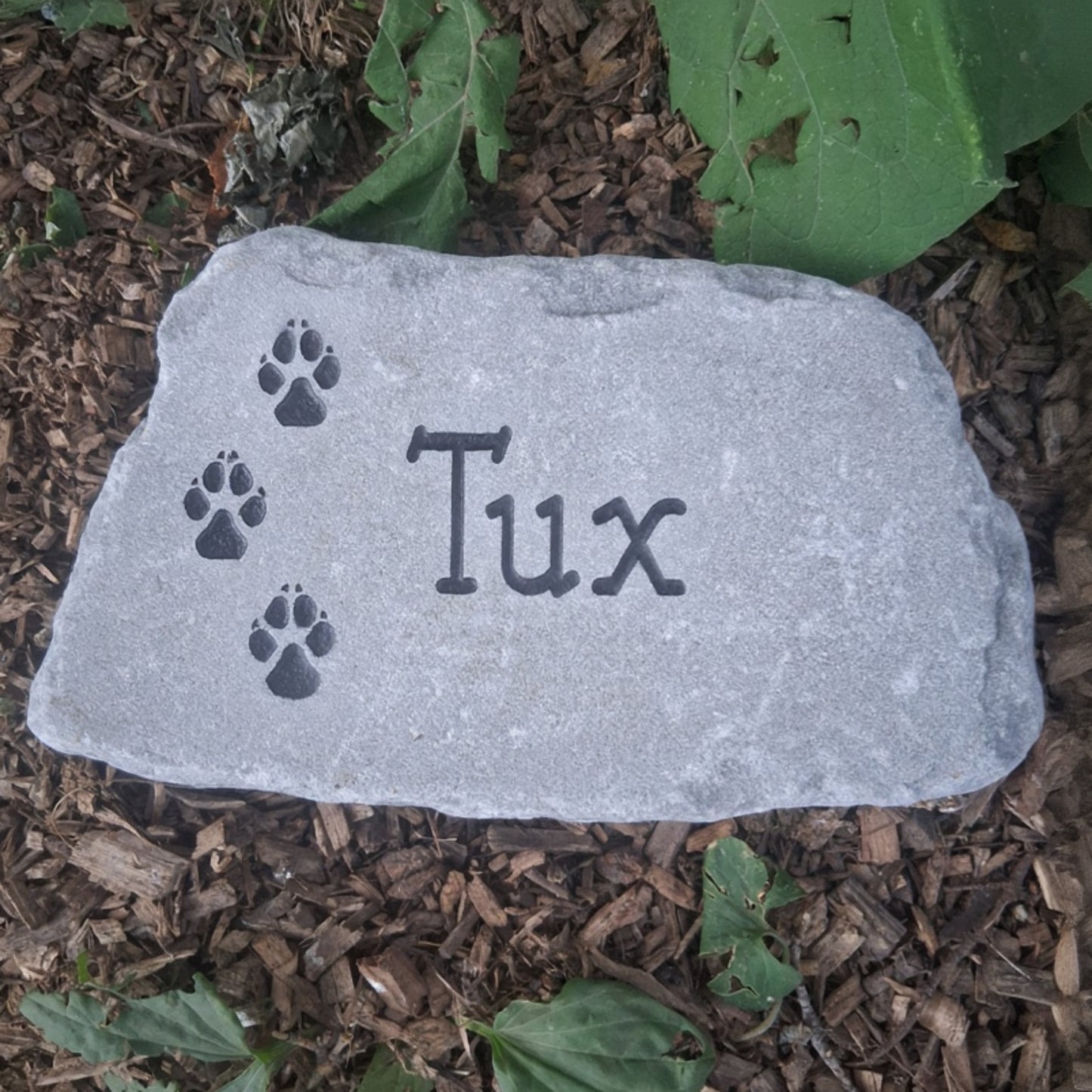 A custom pet memorial garden stone engraved with the name 'Tux' and paw prints, placed on a bed of brown wood chips and green leaves.