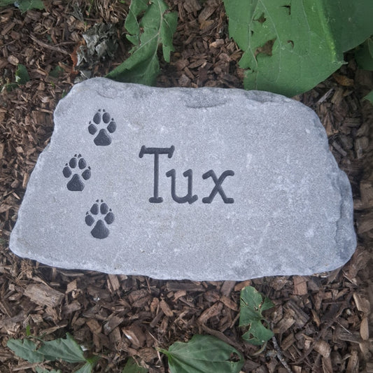A custom pet memorial garden stone engraved with the name 'Tux' and paw prints, placed on a bed of brown wood chips and green leaves.