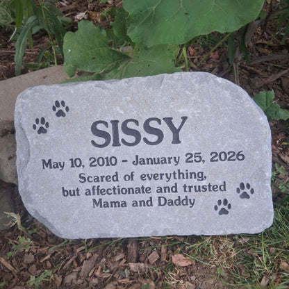 A personalized pet memorial stone engraved with the name 'SISSY', dates, and paw prints, placed outdoors on the ground among grass, mulch, and green leaves.
