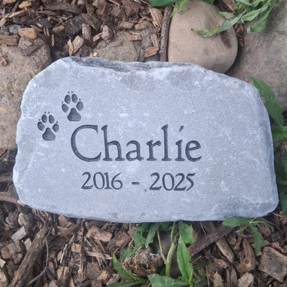 A personalized pet memorial garden stone engraved with the name 'Charlie', the dates '2016 - 2025', and two paw prints, placed on mulch with green plants and other stones in a garden setting.