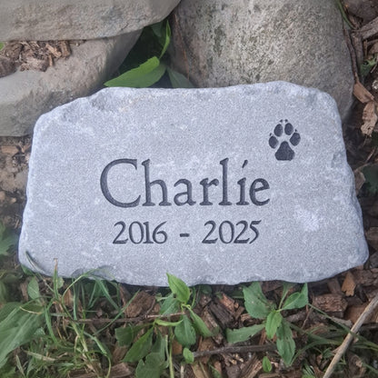 A personalized pet memorial stone engraved with the name 'Charlie', the date '2016 - 2025', and a black paw print, placed on a bed of mulch and green plants.