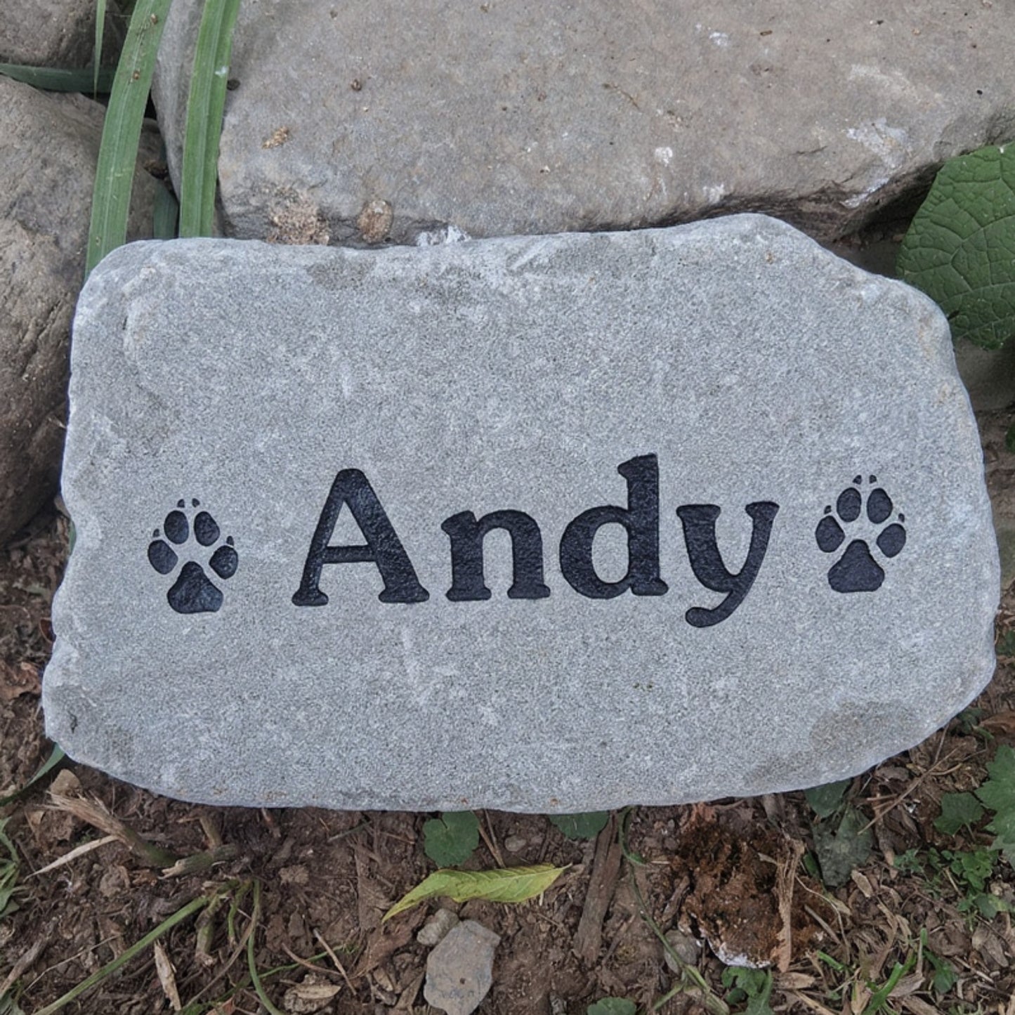 A gray pet memorial garden stone engraved with the name 'Andy' and paw prints, placed outdoors on soil and grass.