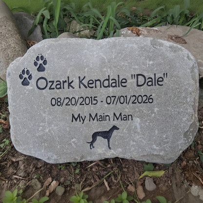 A personalized pet memorial stone engraved with the name 'Ozark Kendale "Dale"', dates '08/20/2015 - 07/01/2026', the phrase 'My Main Man', paw prints, and a dog silhouette, placed outdoors on the ground among grass and soil.