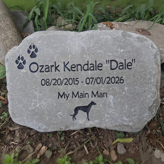 A personalized pet memorial stone engraved with the name 'Ozark Kendale "Dale"', dates '08/20/2015 - 07/01/2026', the phrase 'My Main Man', paw prints, and a dog silhouette, placed outdoors on the ground among grass and soil.