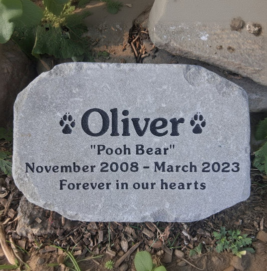 A personalized pet memorial stone engraved with the name 'Oliver', the nickname 'Pooh Bear', dates 'November 2008 - March 2023', and the phrase 'Forever in our hearts'. The stone is photographed outdoors on a bed of mulch with green plants and rocks in the background.
