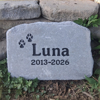 A personalized gray pet memorial stone engraved with the name 'Luna', dates '2013-2026', and two paw prints, placed outdoors on mulch and grass in front of stacked garden stones.