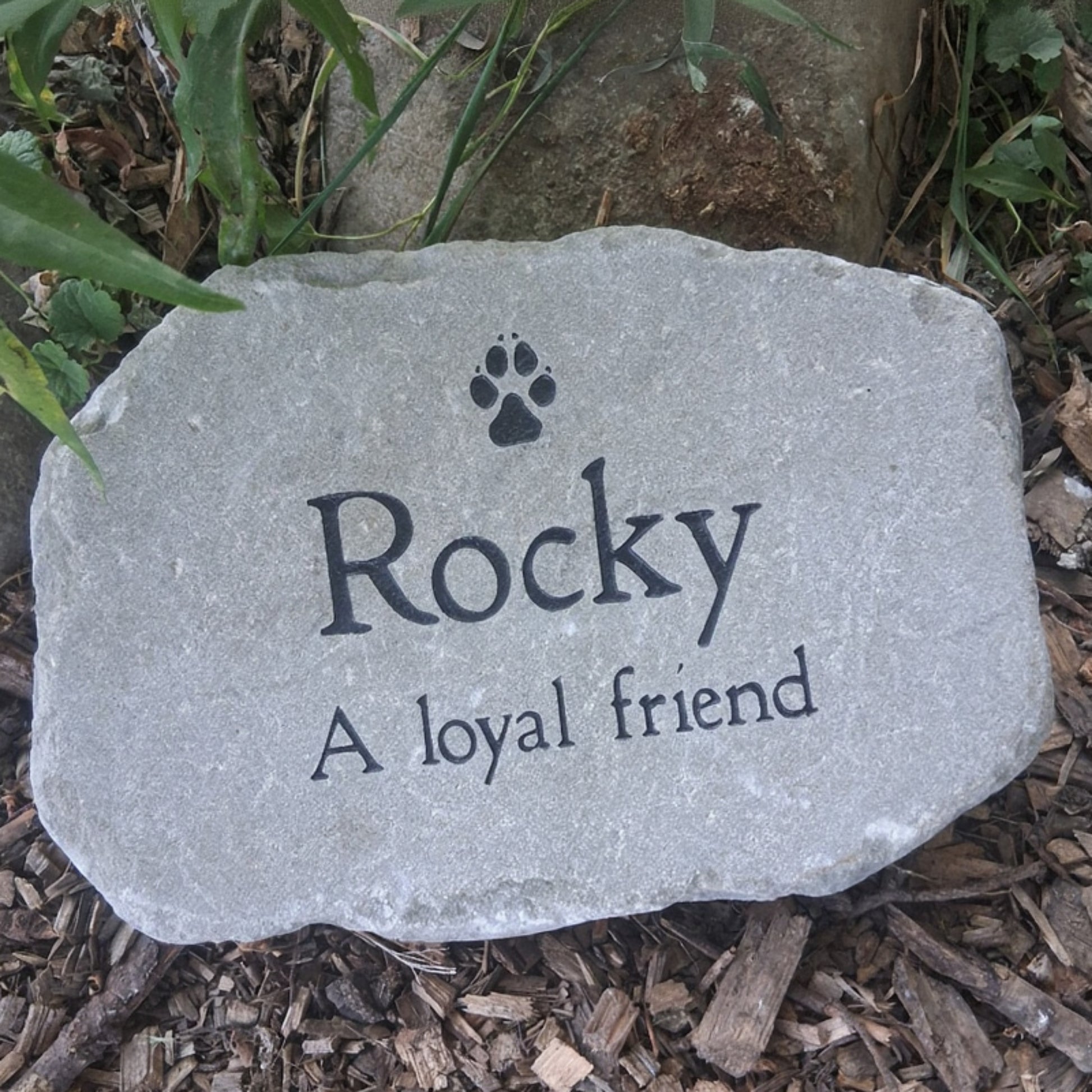 A personalized brown pet memorial garden stone engraved with a paw print and the text 'Rocky A loyal friend', placed on a bed of brown wood chips and green plants.