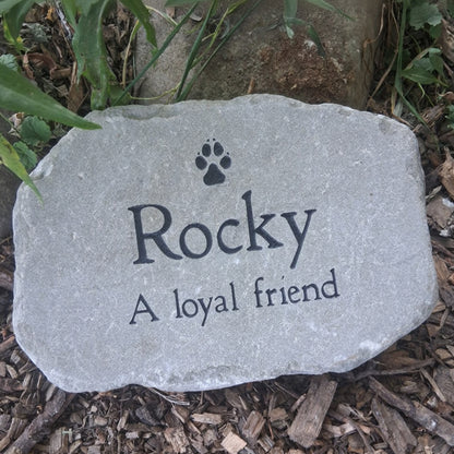 A personalized brown pet memorial garden stone engraved with a paw print and the text 'Rocky A loyal friend', placed on a bed of brown wood chips and green plants.