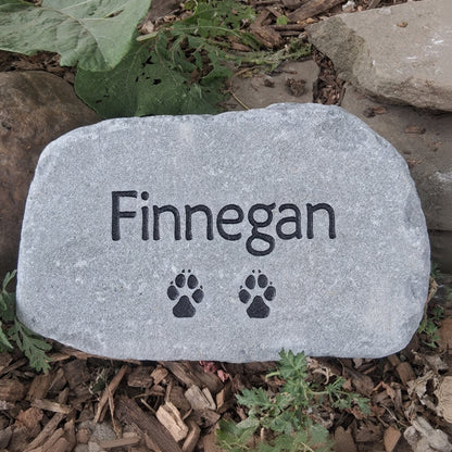 A personalized gray pet memorial garden stone engraved with the name 'Finnegan' and two black paw prints, placed outdoors on a bed of mulch and small plants.