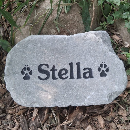 A personalized brown stone pet memorial marker engraved with the name 'Stella' and paw prints, placed on a bed of mulch and green plants outdoors.
