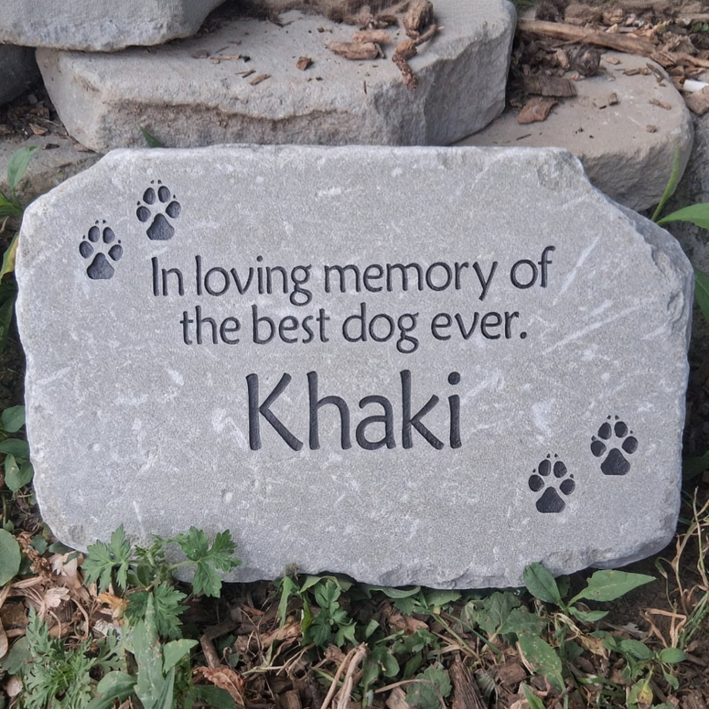 A personalized gray stone pet memorial plaque engraved with the text 'In loving memory of the best dog ever. Khaki' and four paw prints, placed outdoors on the ground among green plants and dried leaves.
