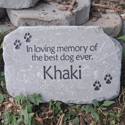 A personalized gray stone pet memorial plaque engraved with the text 'In loving memory of the best dog ever. Khaki' and four paw prints, placed outdoors on the ground among green plants and dried leaves.
