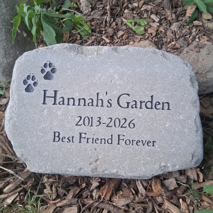 A personalized pet memorial garden stone engraved with 'Hannah's Garden', the dates '2013-2026', and 'Best Friend Forever', along with two paw prints, placed on a bed of mulch and green plants.