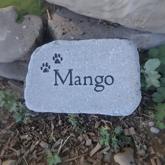 A custom pet memorial garden stone engraved with the name 'Mango' and paw prints, placed outdoors on a stone path next to wood chips and green plants.