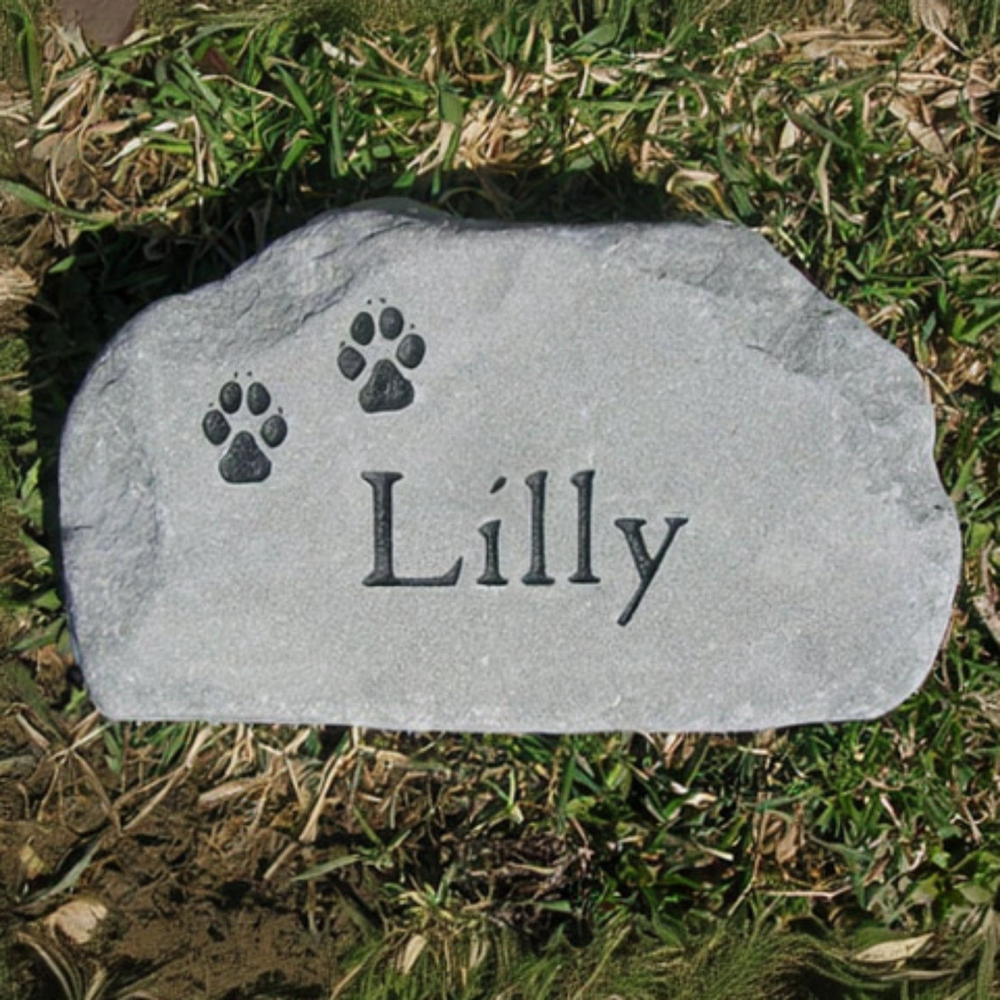A personalized gray pet grave stone engraved with the name 'Lilly' and two paw prints, placed on a patch of green and dry grass lawn.
