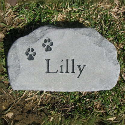 A personalized gray pet grave stone engraved with the name 'Lilly' and two paw prints, placed on a patch of green and dry grass lawn.