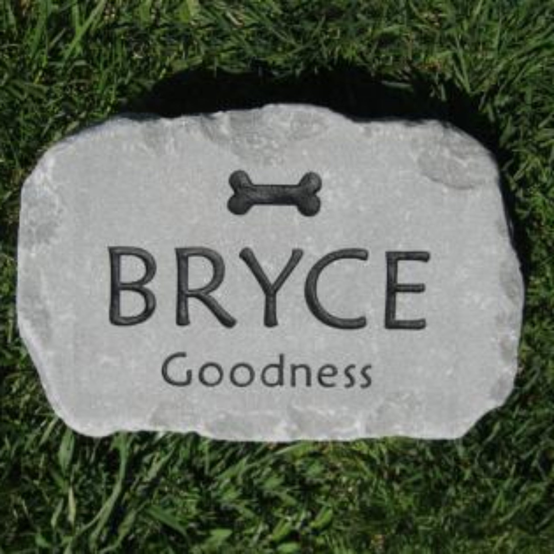 A personalized pet grave stone shaped like a natural rock, engraved with a bone icon, the name 'BRYCE', and 'Goodness' on a green grass background.