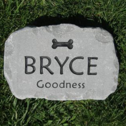A personalized pet grave stone shaped like a natural rock, engraved with a bone icon, the name 'BRYCE', and 'Goodness' on a green grass background.