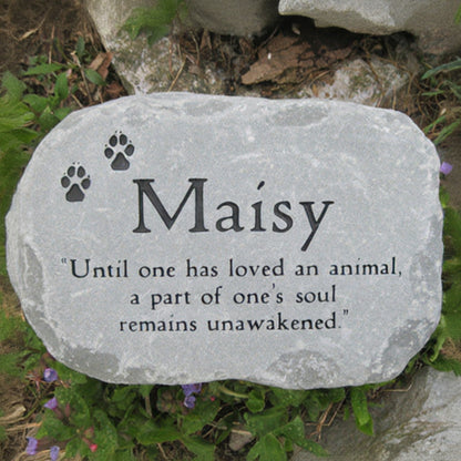 A personalized pet memorial garden stone engraved with the name 'Maisy', two paw prints, and a quote, placed outdoors among grass and small purple flowers.