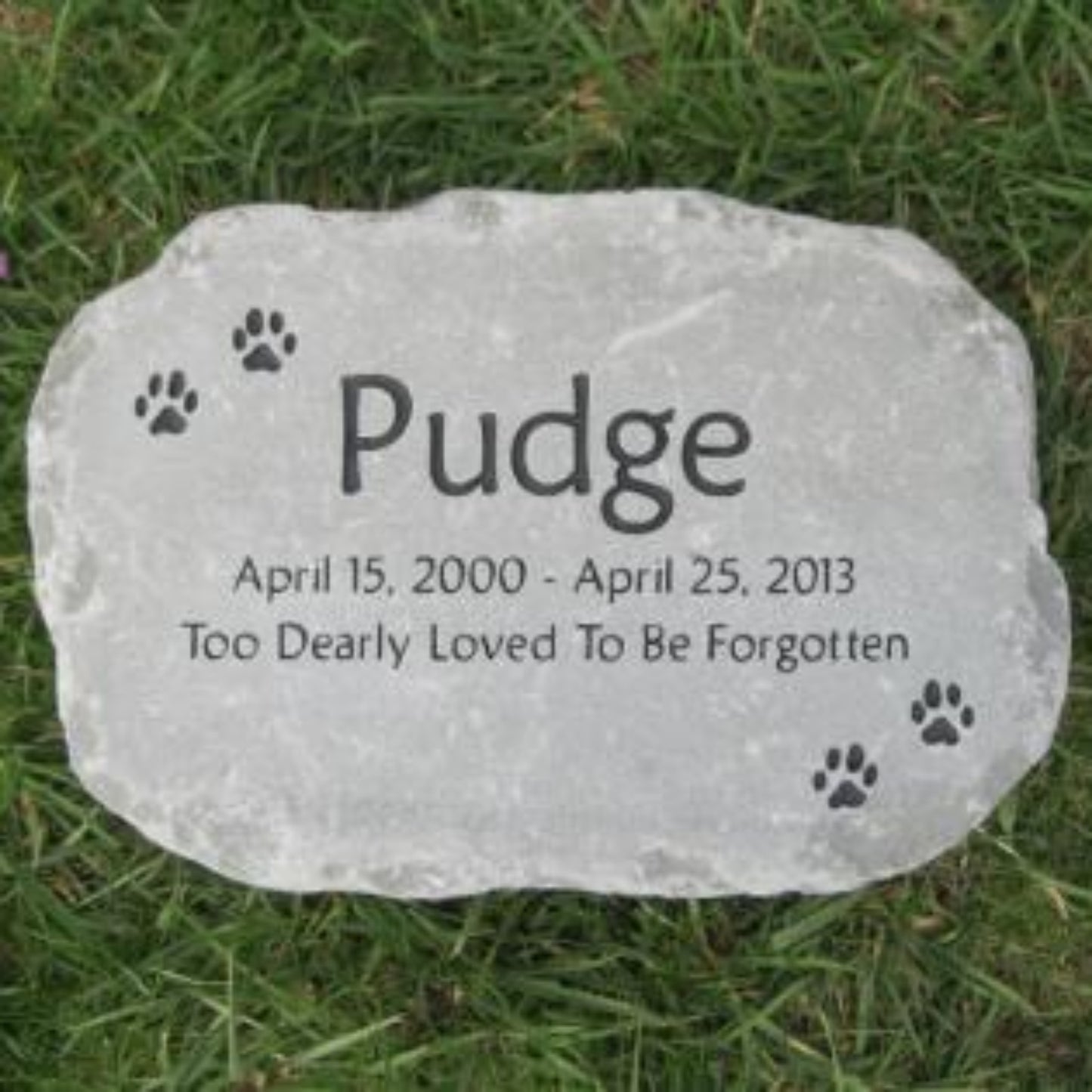A personalized pet memorial stone engraved with the name 'Pudge', dates 'April 15, 2000 - April 25, 2013', and the phrase 'Too Dearly Loved To Be Forgotten', placed on green grass with small paw prints engraved around the border.