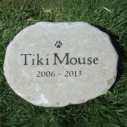 A round light brown memorial pet stone engraved with a paw print, the name 'Tíki Mouse,' and the years '2006 - 2013,' placed on green grass.