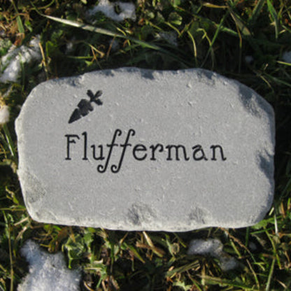 A custom engraved garden stone for a bunny featuring the name 'Flufferman' in a decorative font with a small carrot silhouette above it, placed on grass with patches of snow.