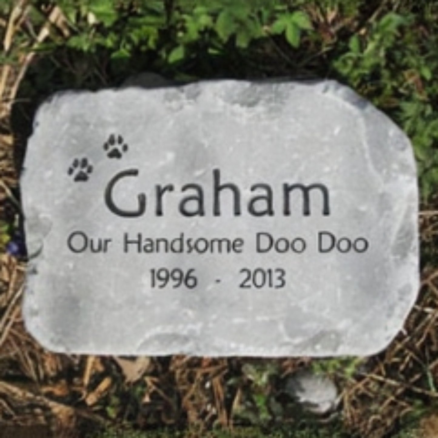 A personalized pet grave stone with engraved text reading 'Graham Our Handsome Doo Doo 1996 - 2013' and small paw prints, placed outdoors on mulch and greenery.