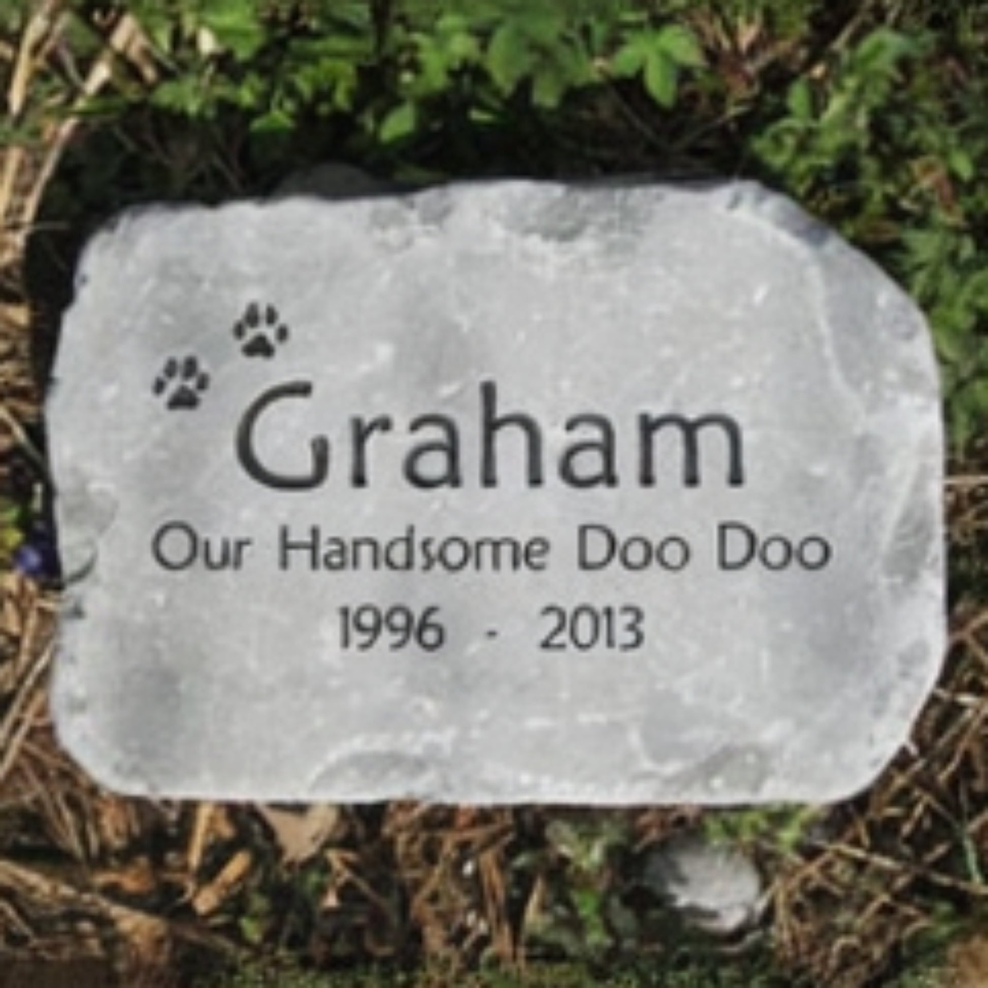 A personalized pet grave stone with engraved text reading 'Graham Our Handsome Doo Doo 1996 - 2013' and small paw prints, placed outdoors on mulch and greenery.
