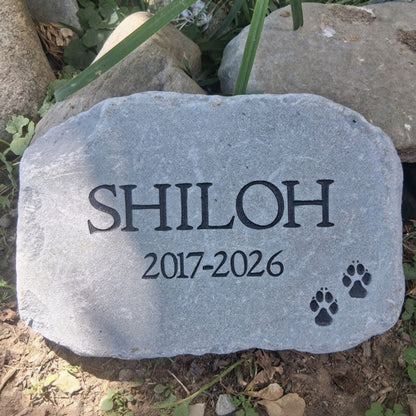 A personalized gray pet memorial stone engraved with the name 'SHILOH', the years '2017-2026', and two black paw prints, placed outdoors on soil and grass.