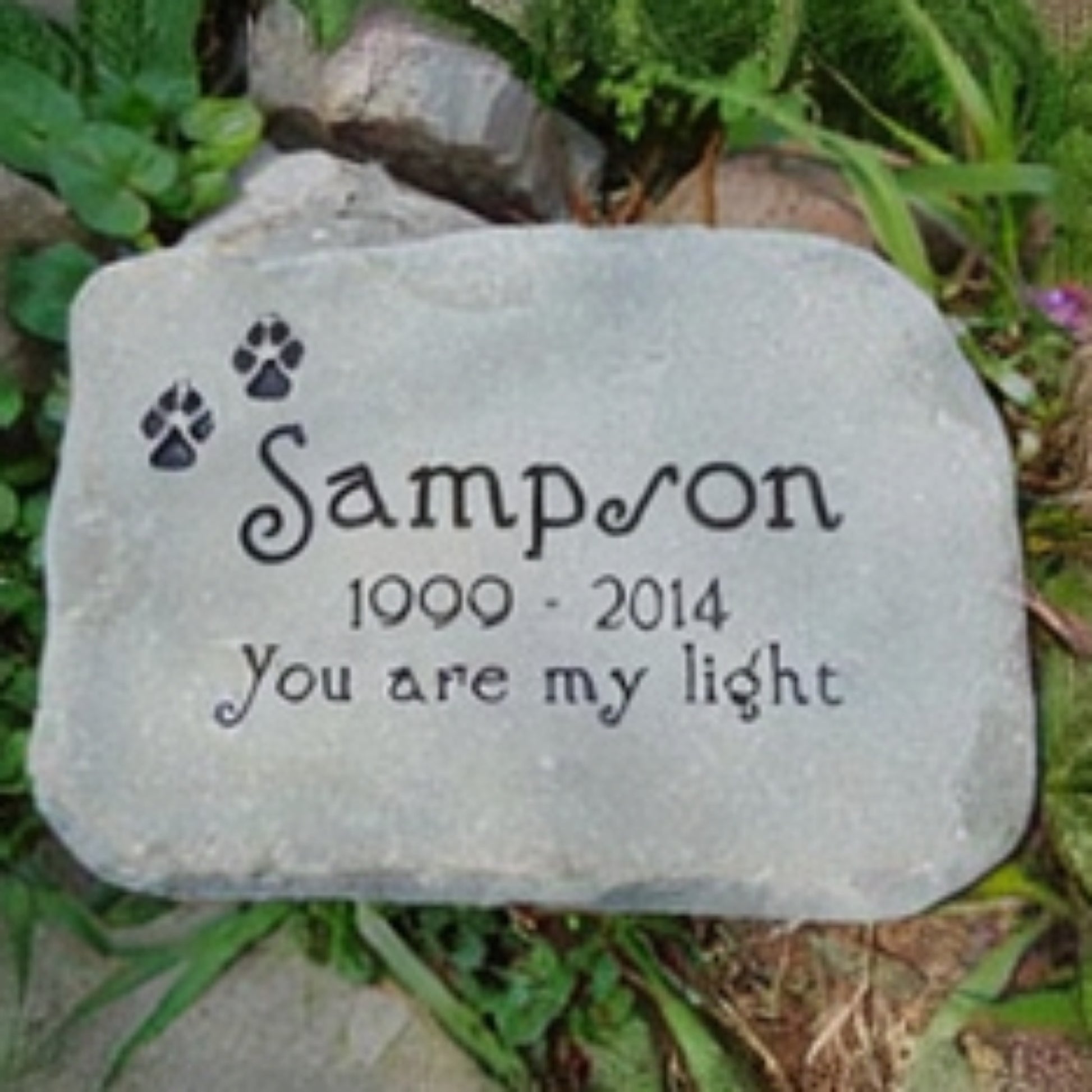 A personalized gray memorial garden stone with engraved black text reading 'Sampson 1990 - 2014 You are my light' and two paw prints, photographed outdoors among grass and rocks.