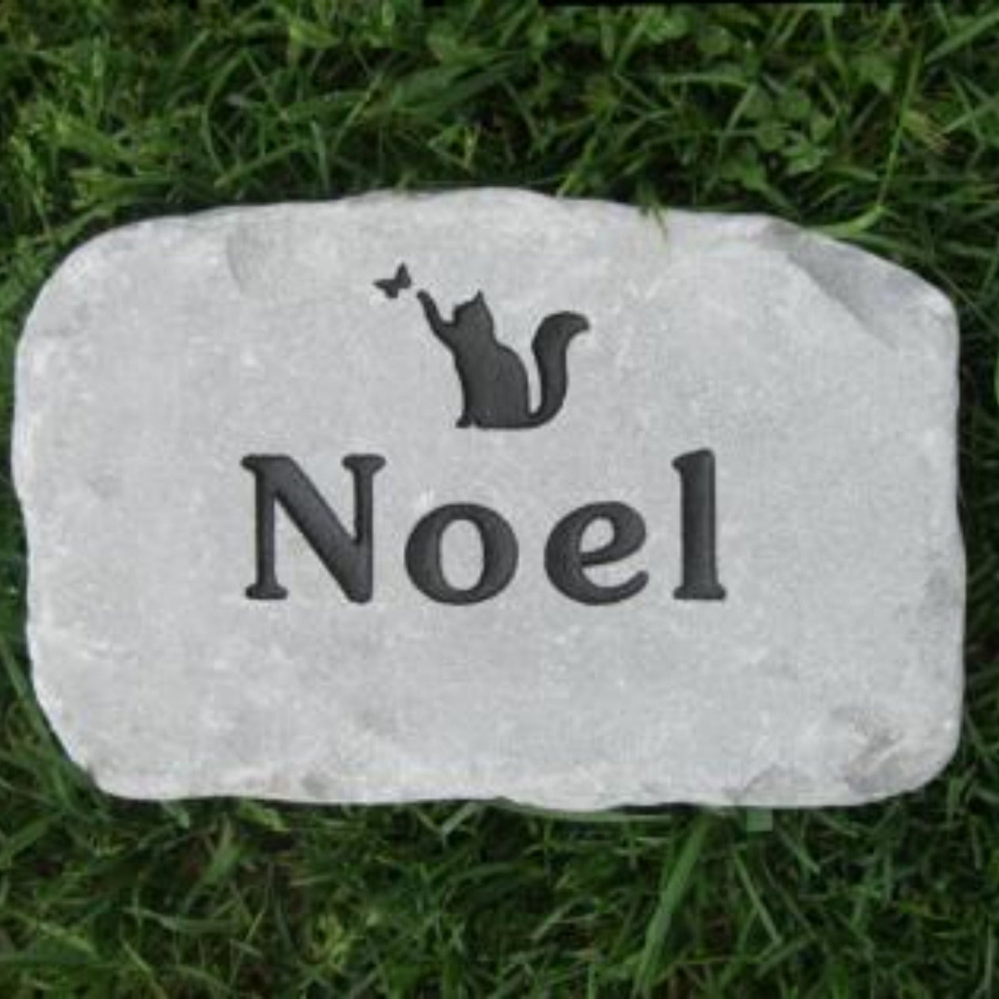 A personalized light gray stone grave marker engraved with a black cat silhouette and the name 'Noel', placed on green grass.