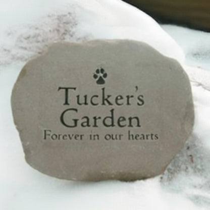 A custom pet memorial garden stone with the text 'Tucker's Garden' and 'Forever in our hearts' engraved, featuring a small paw print icon, shown resting in the snow.