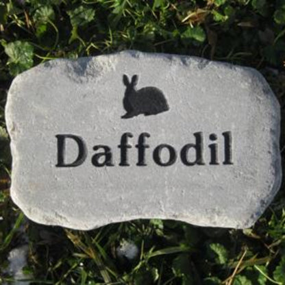 A light gray stone garden marker engraved with a black rabbit silhouette and the word 'Daffodil', placed on green grass with small patches of snow.