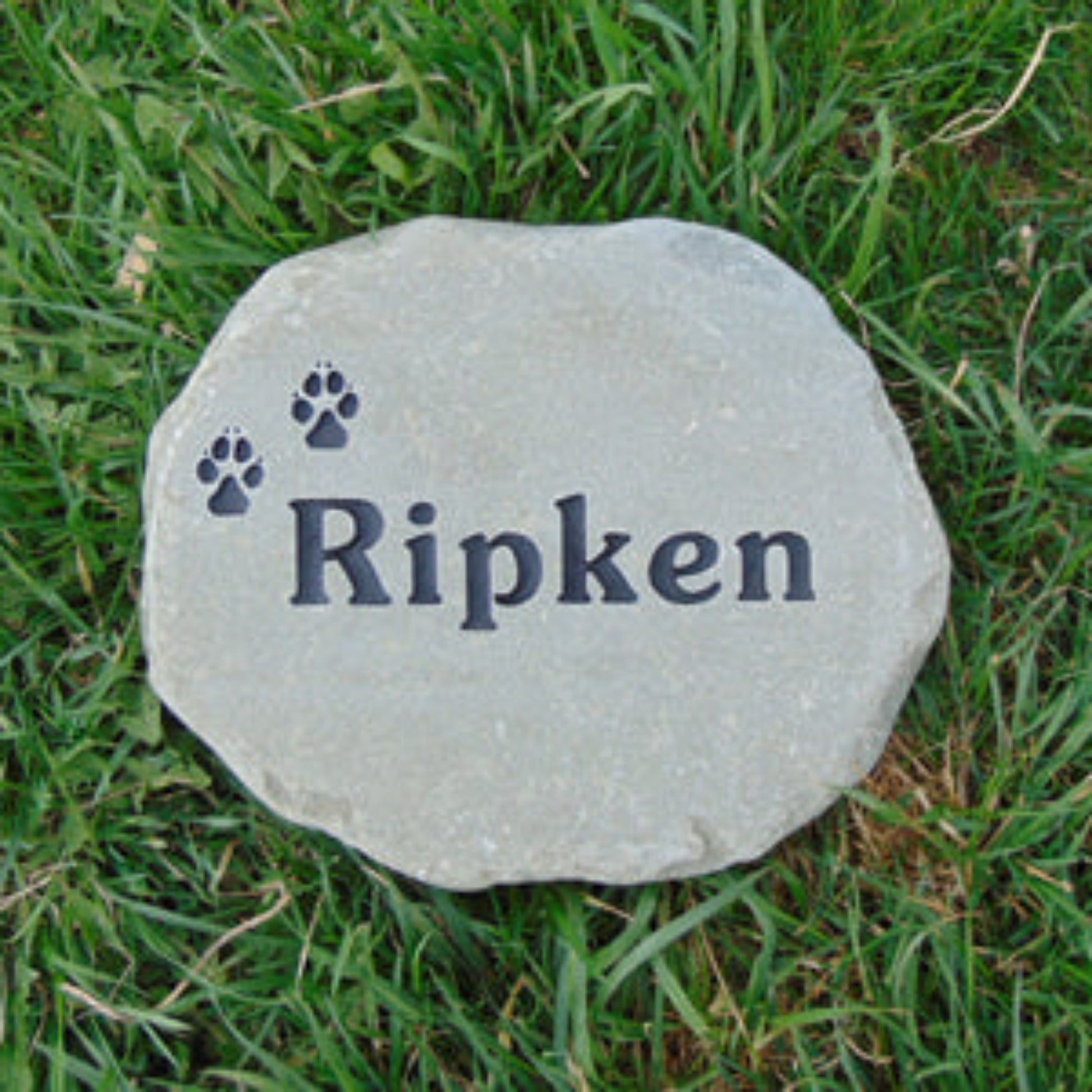 A round bron stone pet memorial marker engraved with the name 'Ripken' and two paw prints, placed on green grass.